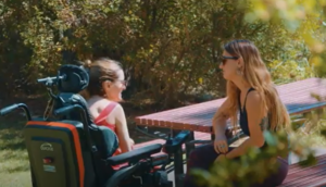 A person in a wheelchair talking to a person sitting on a park bench at a table.